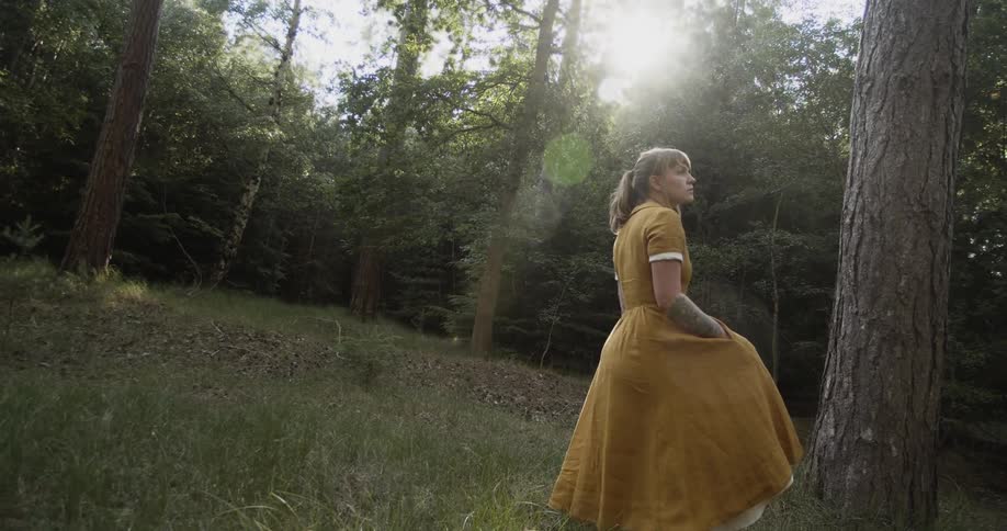 Woman stands in a forest at daytime with her hand in her dress pocket swaying from side to side