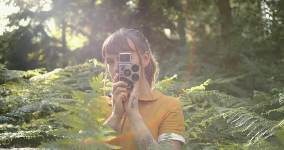 A flare of sunlight shines behind a woman with a wind up camera pointing at the camera standing in forest at daytime