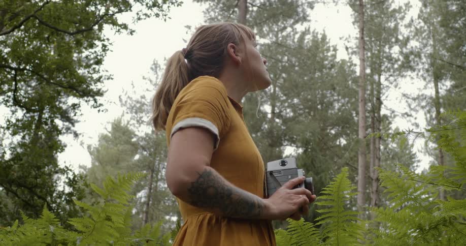 Woman standing in a forest holds a camera while looking up around her in wonder at daytime