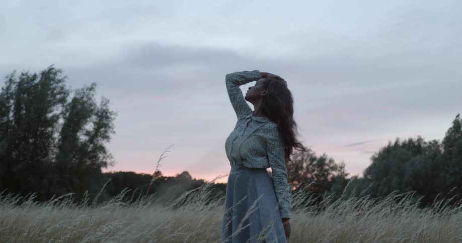 A woman standing in a grassy field with trees the sky and clouds in the background