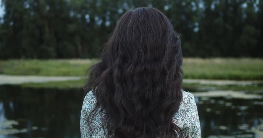 A woman with dark hair slowly walks outdoors towards a pond