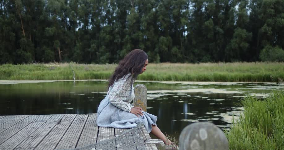 Young woman wearing a dress sitting in a pier on a lake in the forest