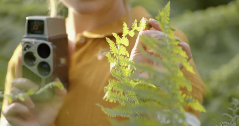 A woman standing outdoors while surrounded by vegetation examines a branch of a tall plant while holding a camera on a sunny day