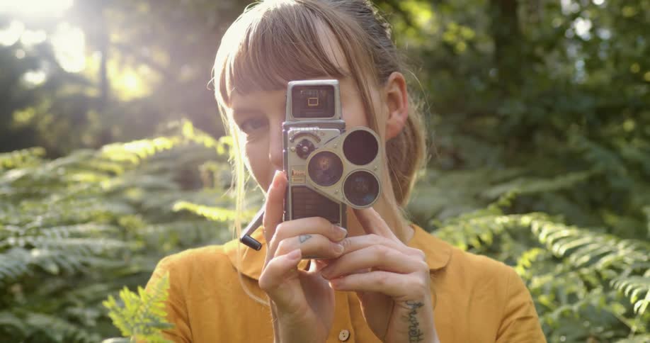 A young woman holds a vintage camera up to her eye while posing for the camera