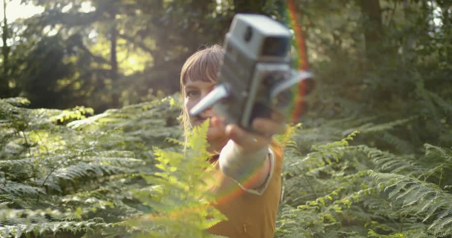 Young woman wearing a yellow dress holds a vintage film camera in the forest at sunset