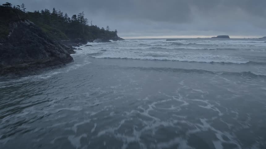 Aerial pushes over shoreline wave breaks against rocky coast and stormy skies