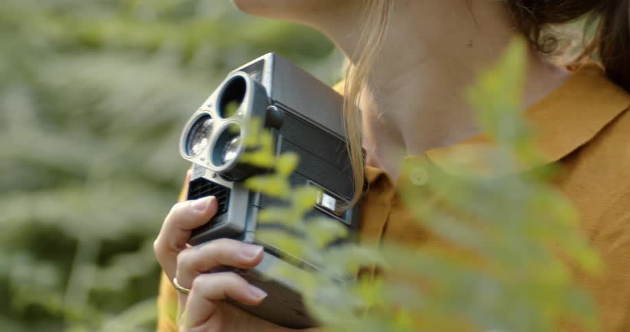 A woman holds a vintage camera as she touches a lush bush in the forest