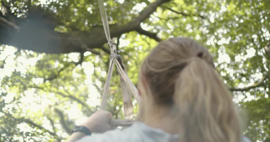 A woman holding on to a swing with trees leaves the sky and clouds in the background