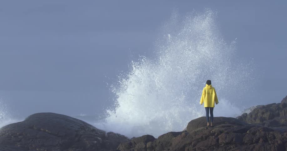 Woman wearing yellow jacket stands on a rocky bank as a huge wave splashes high in front of her
