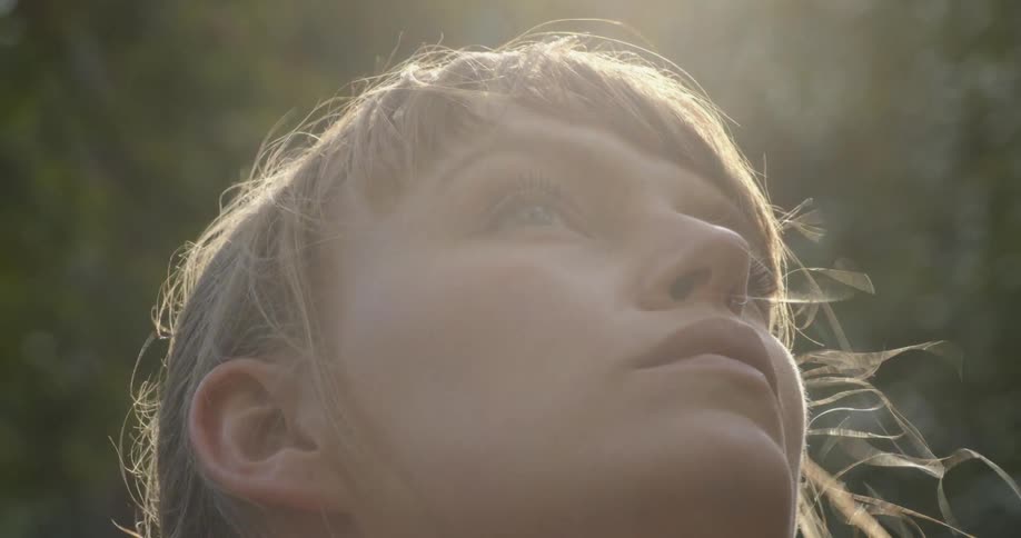 A woman looking upwards with wind blowing through her hair with trees and sunlight in the background