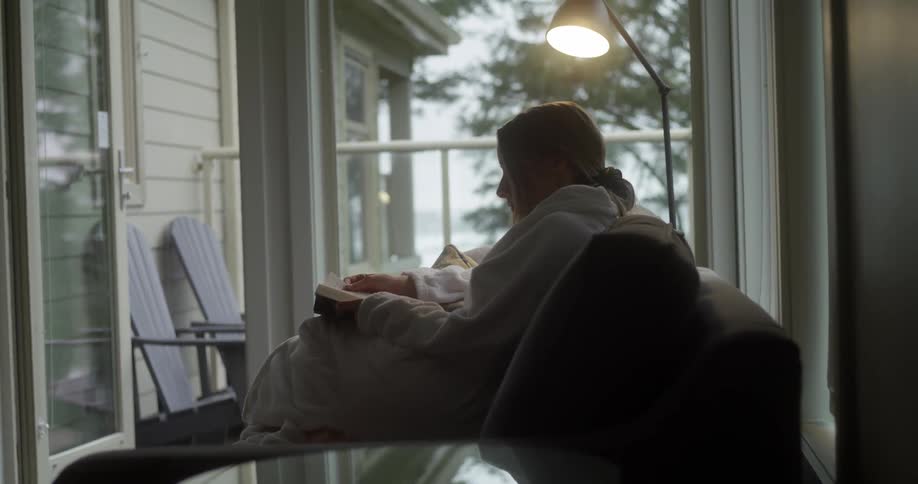 Woman sits alone on couch reading a book next to a balcony