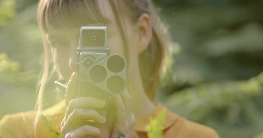 Young woman looking at the camera through the lens of a vintage film camera in the forest