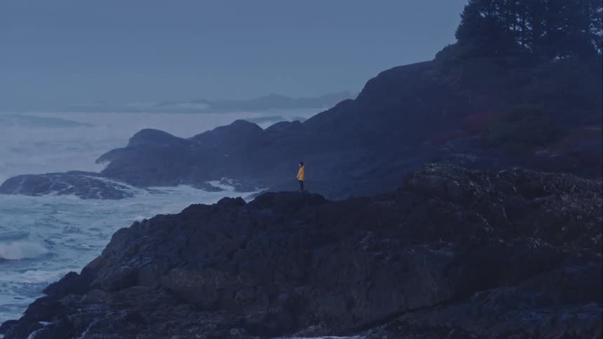 Aerial approaches woman standing on jagged rocky outcrop as waves crash around her