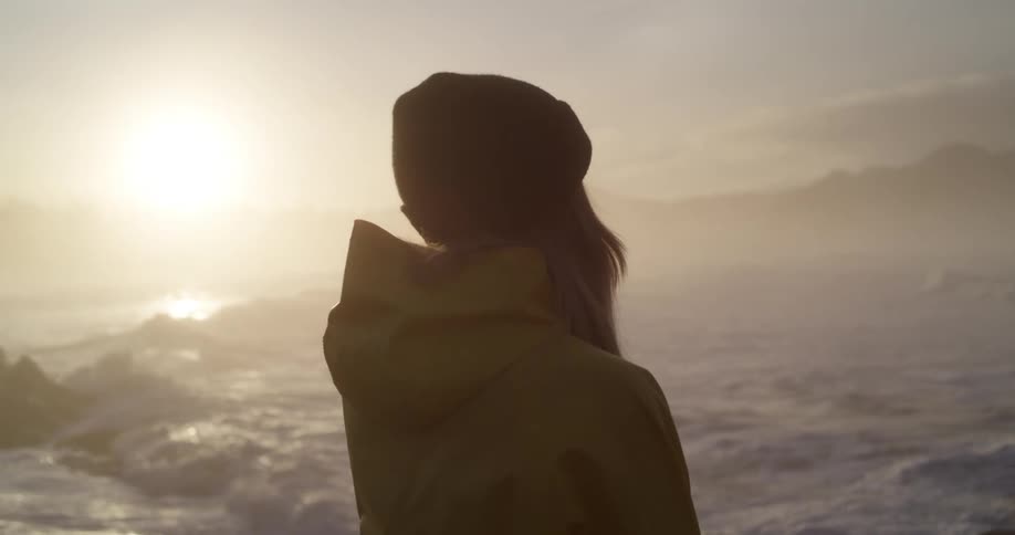 Young blonde woman wearing a bright yellow rain coat looking at the ocean at sunrise