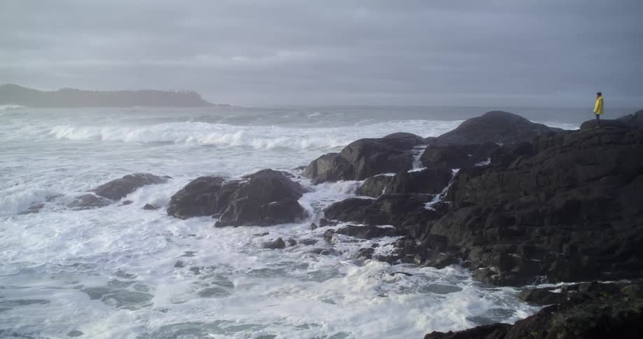 Waves crashing and splashing against coastline rocks as woman wearing a yellow jacket stands on top of the rocks watching