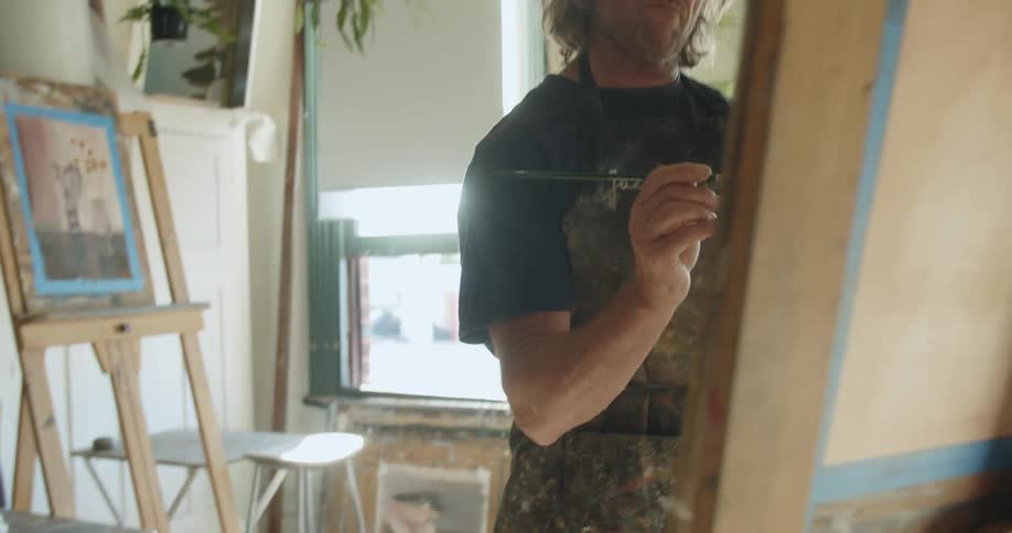 A man painting on an easel in his studio with lots of windows and plants