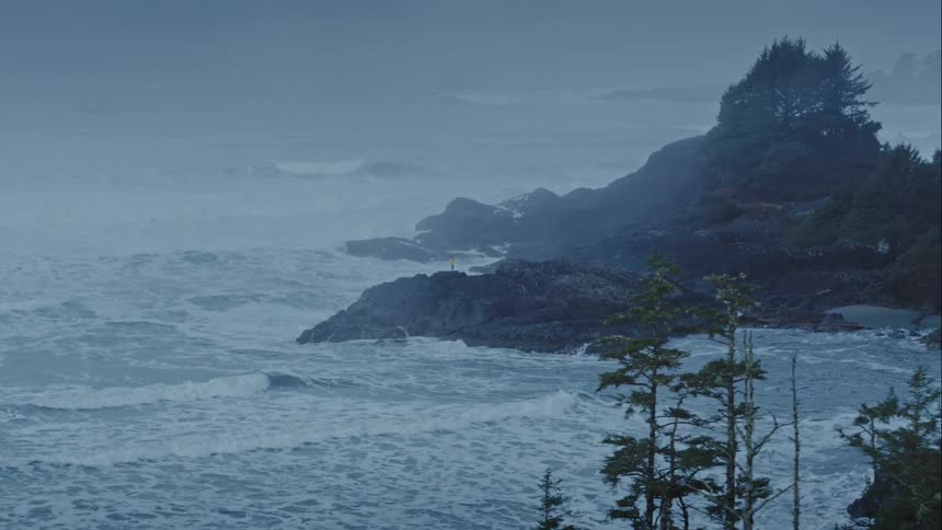 Woman in yellow jacket stands on a rocky outcrop surrounded by stormy ocean waves crashing