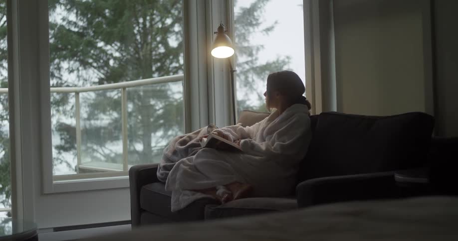 Woman sits on a couch in a living room holding a book as she stares out the window at daytime