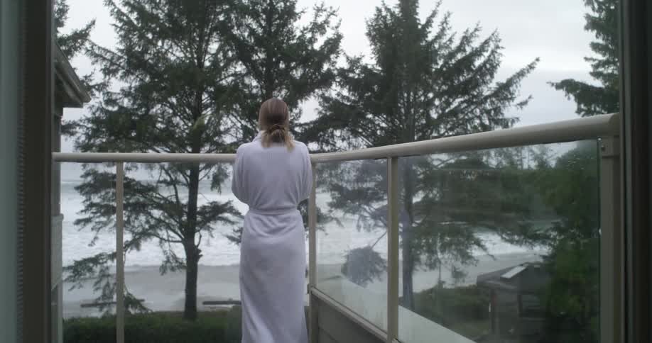 A woman in a white robe stands on a balcony overlooking a beach