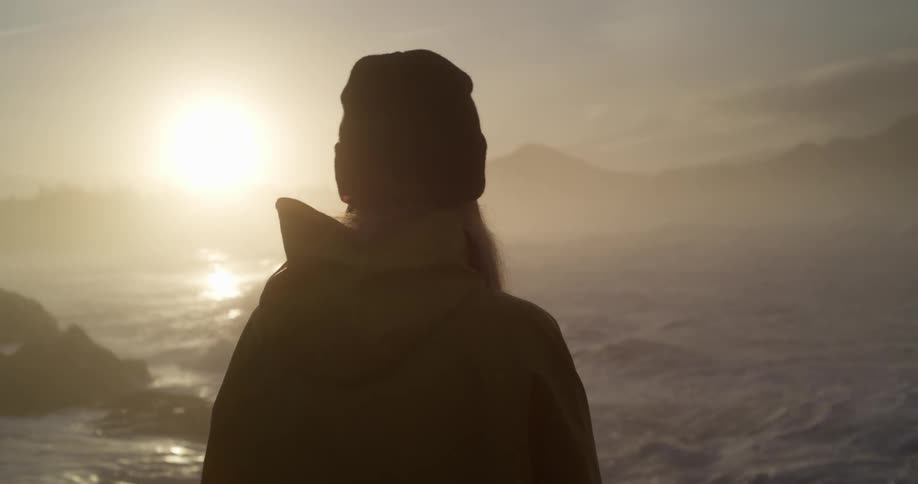 A woman standing in front of an ocean at sunrise as waves crash