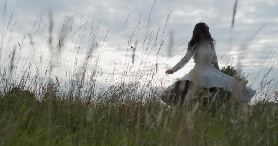 A woman with a dress spinning in a grassy field with trees the sky and clouds in the background