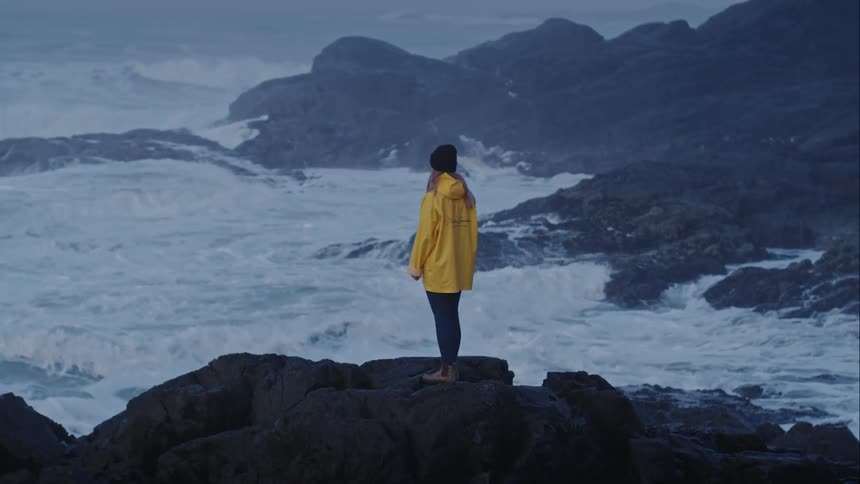 Aerial of woman standing on rocks while stormy ocean waves crash around her