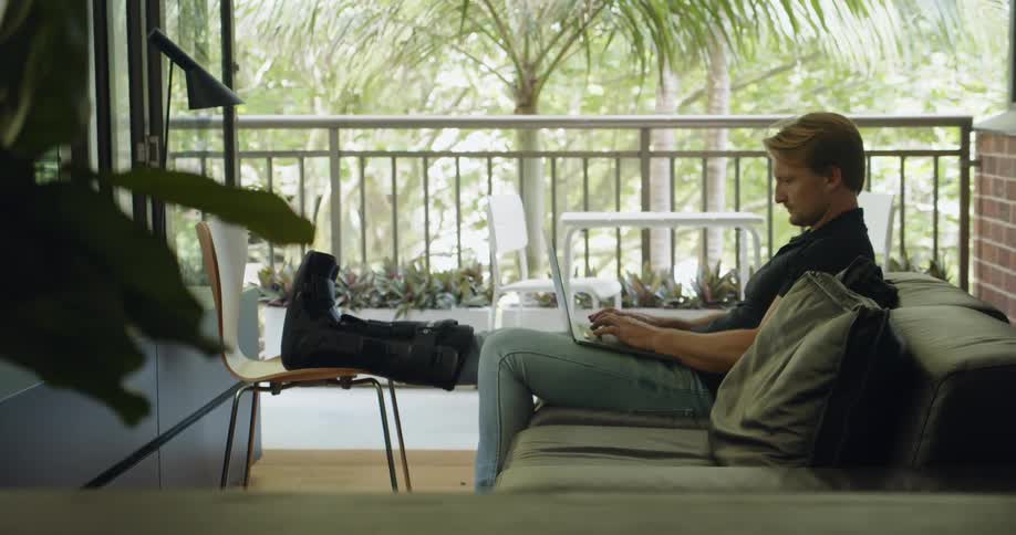 A man types on a laptop sitting on a couch with his casted leg propped on a chair