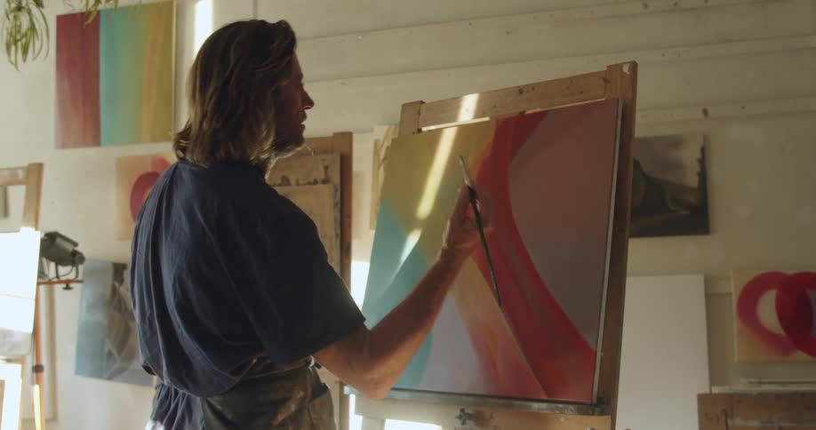 A middle aged man dips a paintbrush into paint on a dish before raising it in preparation to paint on a canvas on an easel