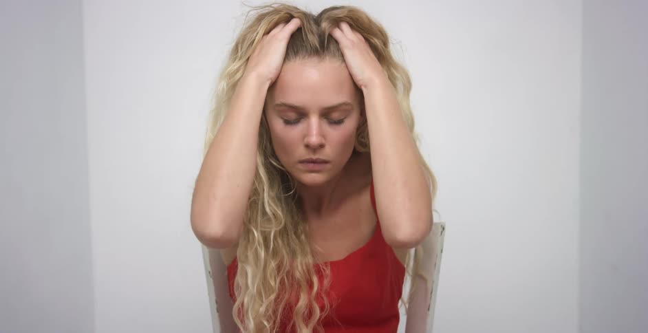 Sad young blonde long haired woman combs her hair with her hands sitting in the corner of a white room