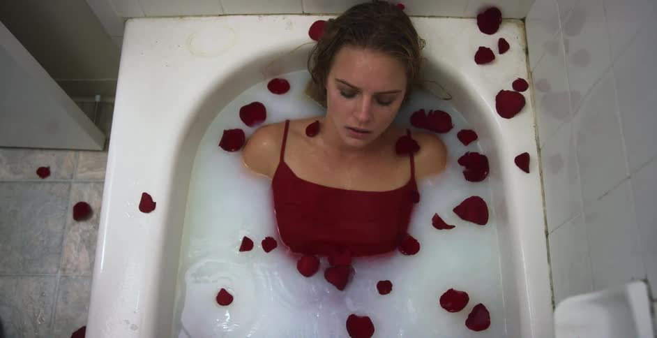 A woman is in a white bathtub with her eyes closed and rose petals dropping down on her