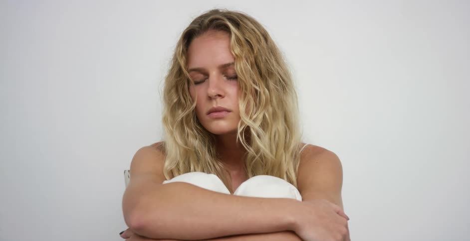 A woman sits against a white wall with her eyes closed as she sways back and forth