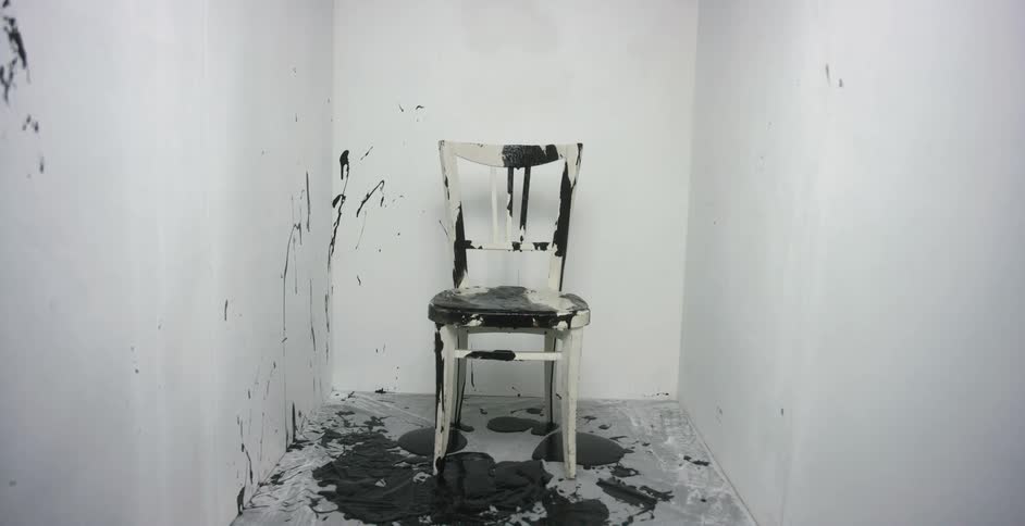 Black paint is splattered onto a white chair sitting at the end of a narrow room with white walls