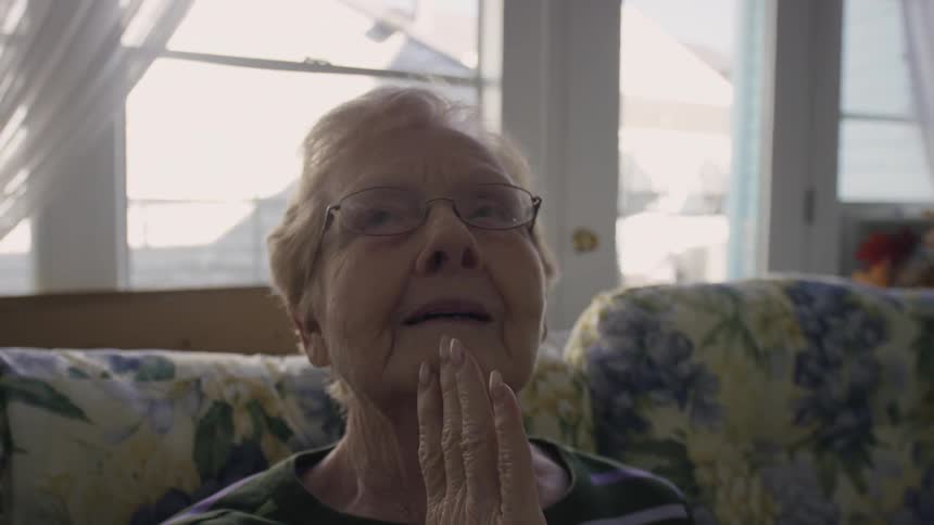An elderly woman rubbing her cheek and smiling as she sits on a couch