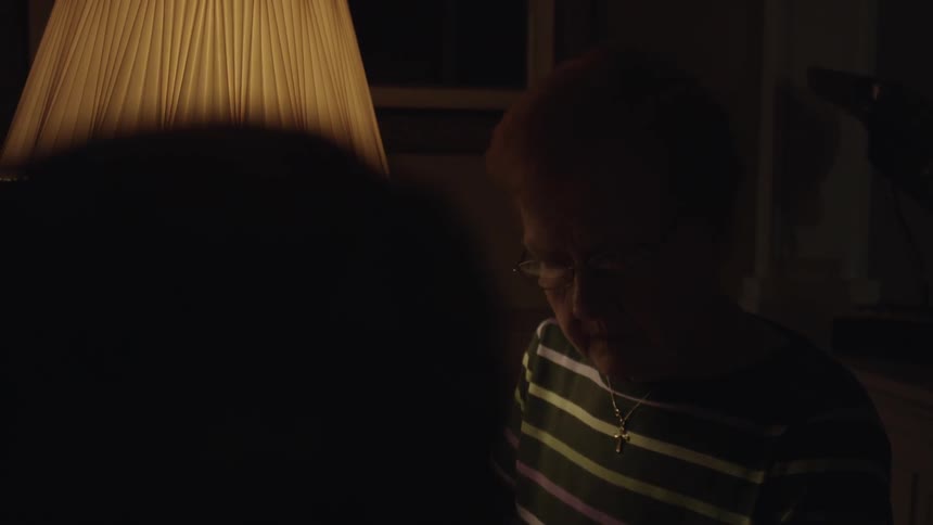 An older woman sits by a lit lamp and interacts with something off camera