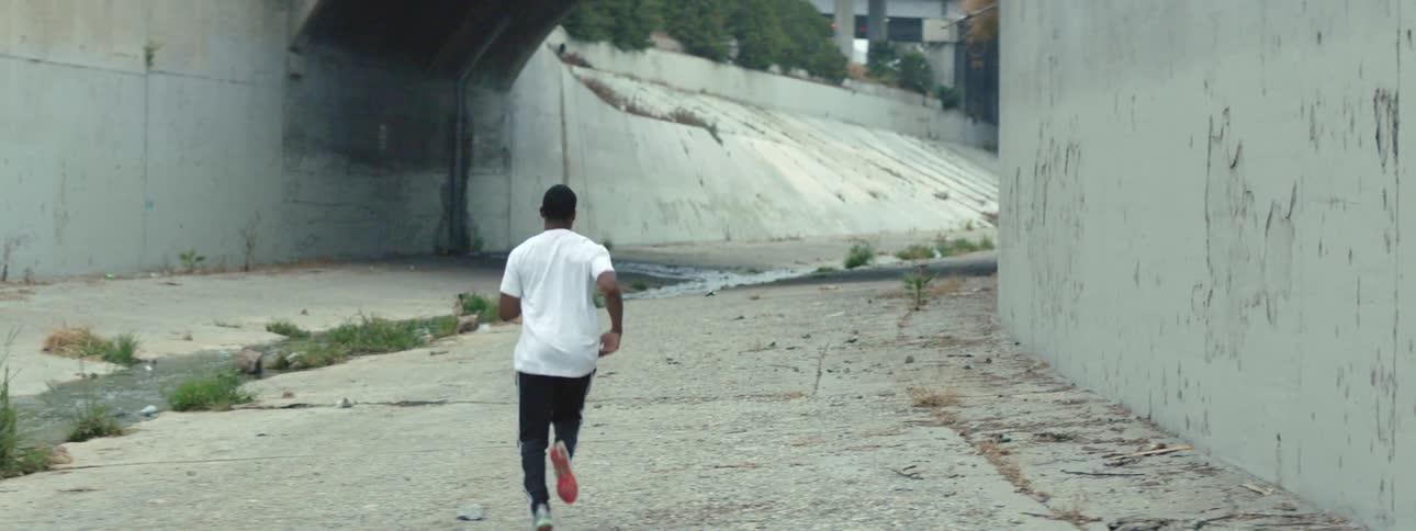 A man jogs down an alley that runs past a large white wall and ramp in a city