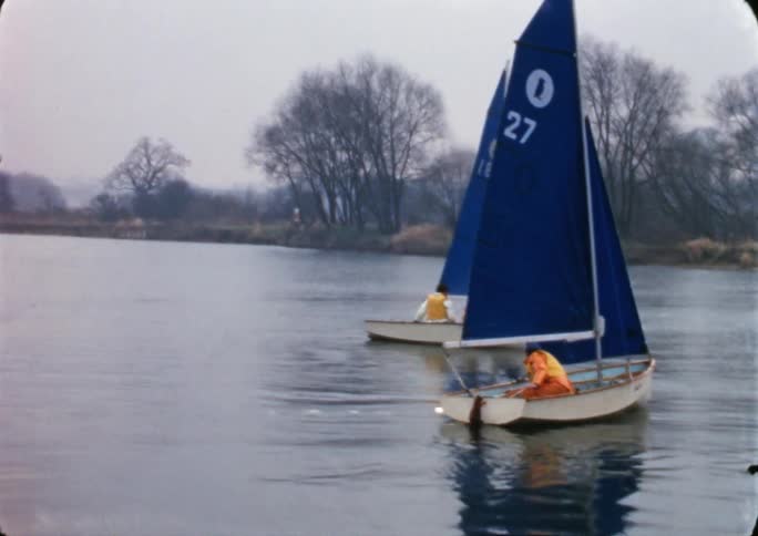 People ride a pair of small sailboats across a lake on a cloudy fall day