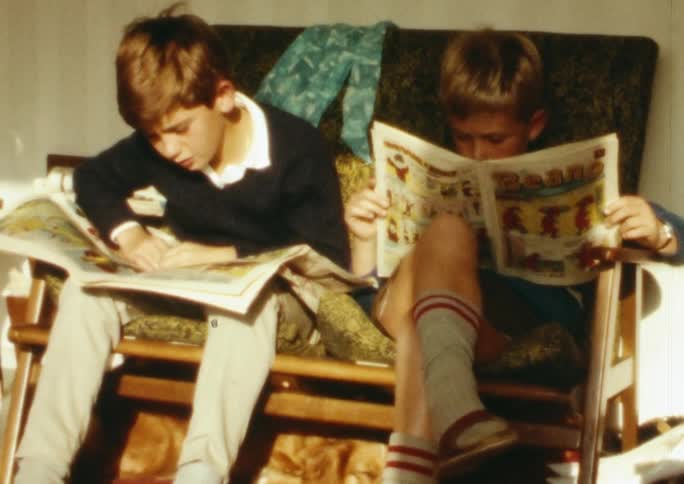 A pair of boys sit together on a small sofa while reading comic books during the day