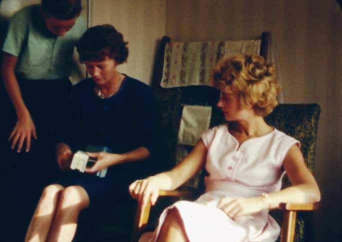 A boy walks over to two women who are sitting down fiddling with a device inside of a room at daytime