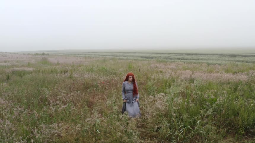 A woman in a dress and boots walking on a grassy field with the sky and clouds in the background