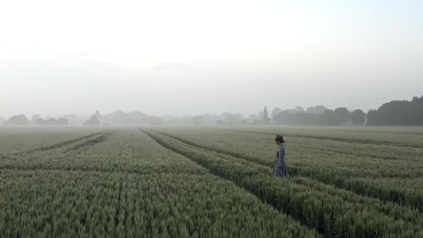 Woman wearing hat and dress is walking through a field of crops with trees in the distance on a hazy day