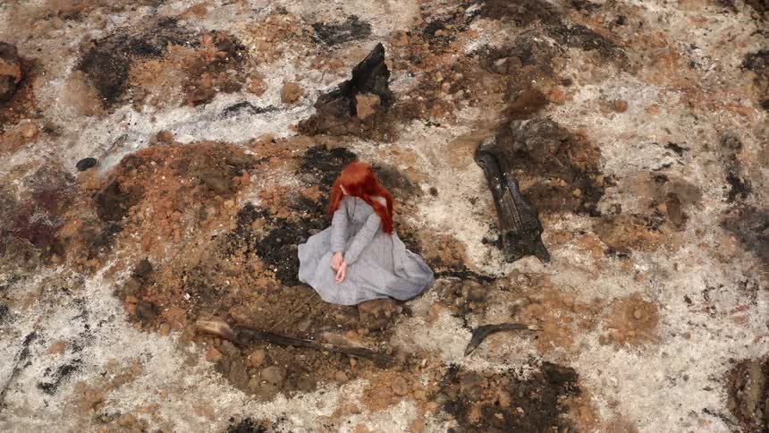Long haired red head young woman wearing a long blue dress sitting over the dry moss on a field