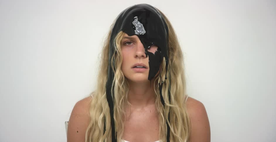 Long haired blonde woman sitting in a white room with black paint smeared over her head and face