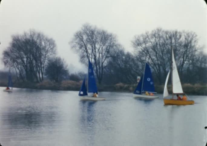 A group of small sailboats sail slowly down a river on a cloudy autumn day