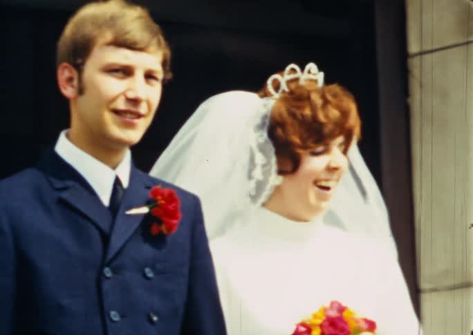 A bridge and groom hold hands while standing in the doorway of a large building as they speak and pose for photographs