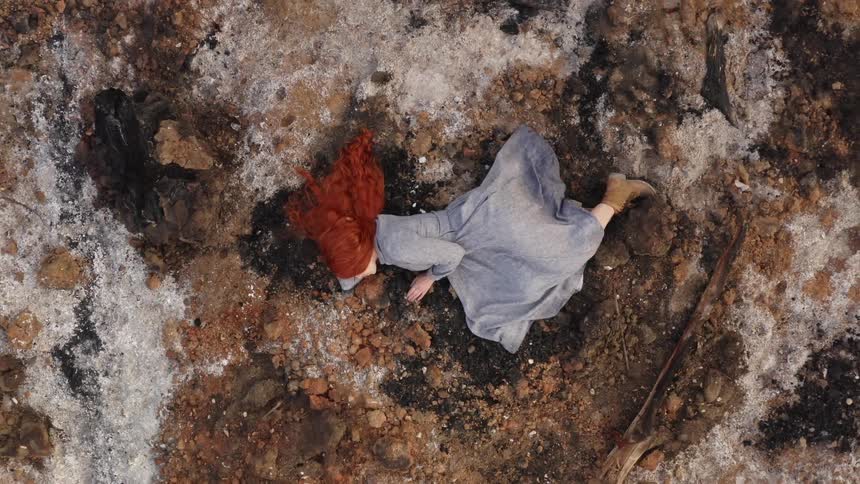 Red headed woman wearing a blue dress lying on her side on the floor outdoors on top of rocks and dirt during the day