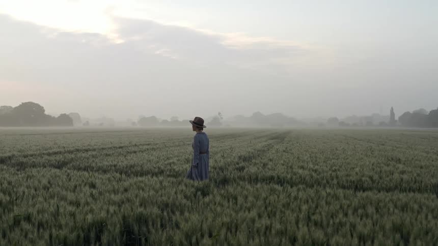 A woman wearing a hat and blue dress walks through a field of crops on an overcast day