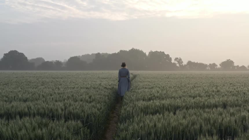 Woman wearing a long blue dress walking on a trail in a field at sunset