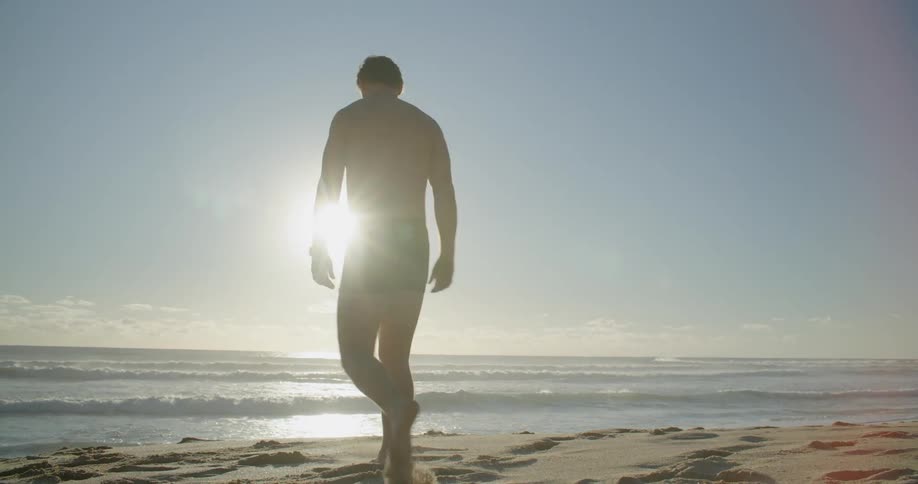 A man wearing swim trunks walks on the beach towards the ocean in bright sunlight