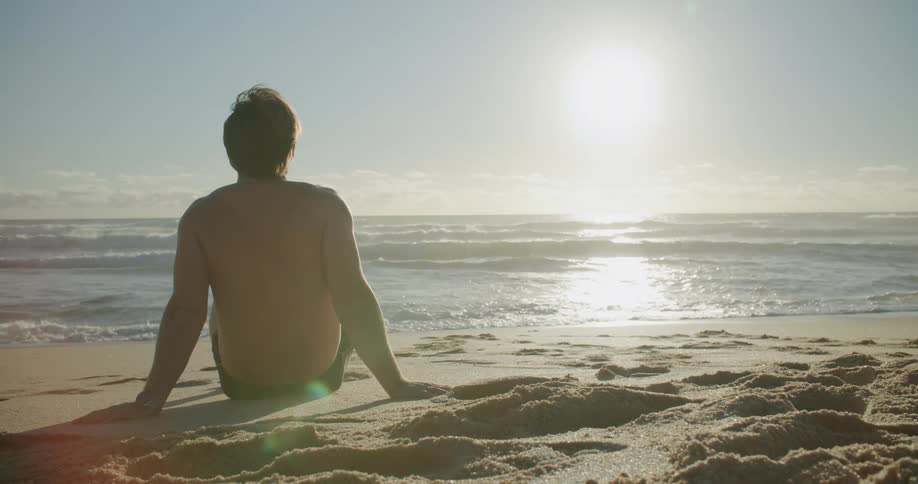 A man sits on a sandy beach and looks out upon the ocean on a sunny day