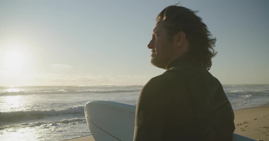 Man holding a surfboard over the sand of a beach next to the ocean at sunset